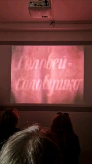 Показывают короткий фильм в небольшом зале музея: проекция кадра на экран и силуэты посетителей в тёмной комнате.