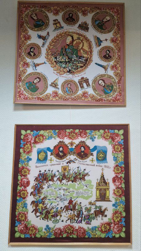 Два оформленных платка в рамках на белой стене музея: разнообразие орнаментов и иллюстраций, показывающих тему платочного искусства.