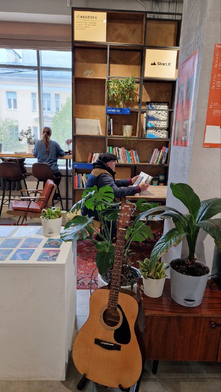Уютный уголок внутри арт‑кластера: книжные полки, стулья, гитара у стойки и горшечные растения, создающие ощущение домашнего пространства.