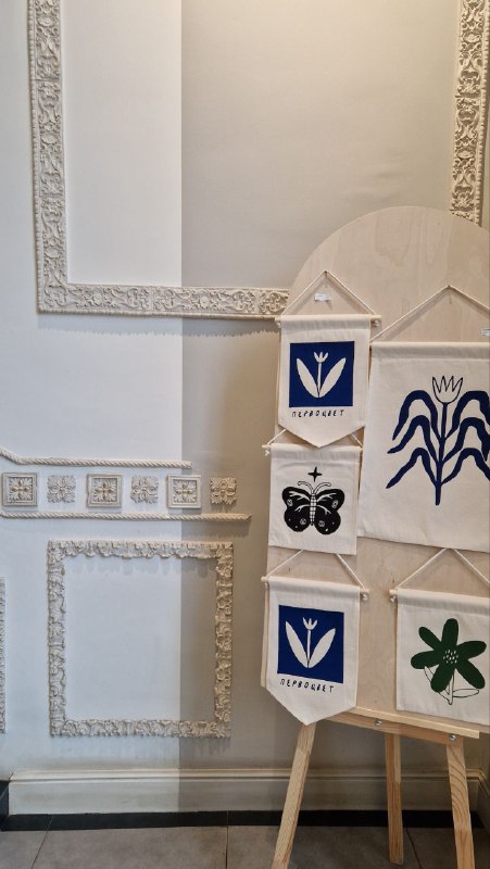 Детали интерьера с оригинальной лепниной и экспозицией на мольберте: карточки и флажки местных мастеров на фоне белой стены.