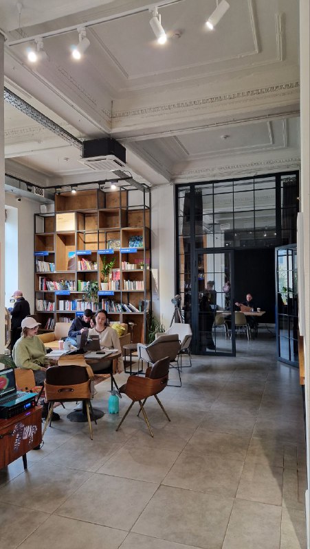 Широкий вид внутреннего зала арт‑кластера: панорамные окна, высокие потолки с лепниной, столы и сидящие посетители в светлом пространстве.