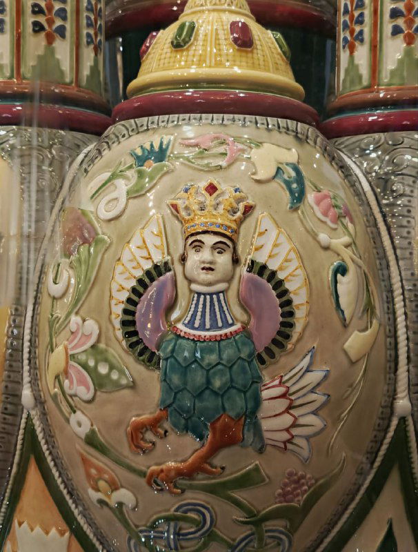 Декоративный фарфоровый или керамический сосуд в экспозиции музея с изображением коронованного крылатого существа и растительным орнаментом.