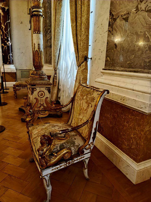 Антикварное кресло с позолоченными элементами и резной отделкой на фоне интерьерных деталей одного из залов дворца.