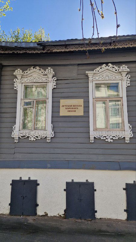 Серый деревянный дом с двумя окнами и белыми резными наличниками, на фасаде табличка с информацией — пример сохранившейся городской застройки.