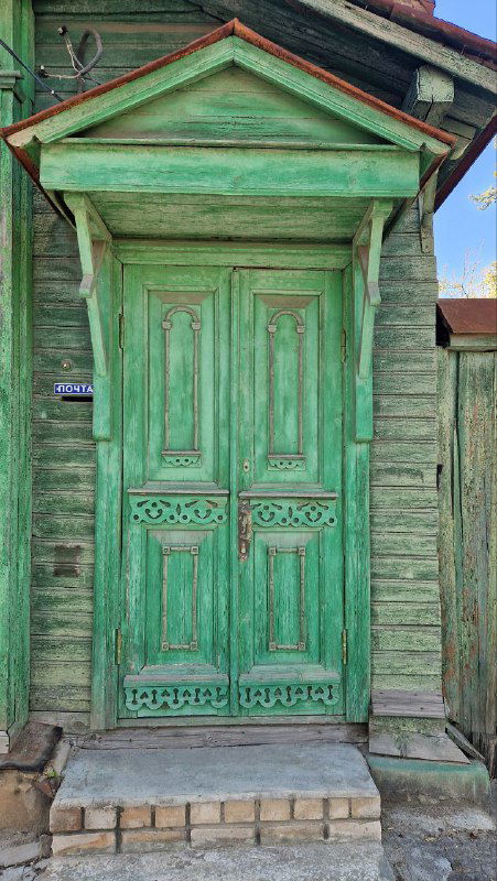 Зелёная двустворчатая деревянная дверь с наличниками и резным декором — типичный вход в жилой дом Затьмачья, фотоснимок в солнечный день.