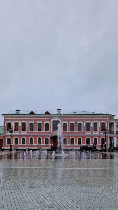 Фасад Императорского путевого дворца в Твери на пасмурном небе; мокрая площадь и фонтан видны перед главным входом и колоннами здания
