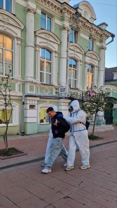 Двое людей в защитных костюмах идут по улице Твери рядом с витриной — возможно уличный перформанс или костюмированное событие в городе