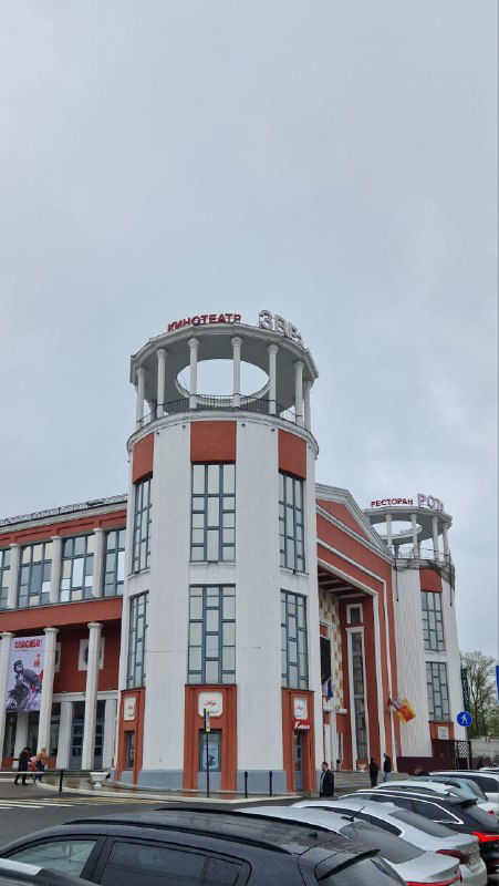 Необычное современное здание с колоннами и башенками в Твери; перед ним парковка и автомобили на городской улице рядом с входом