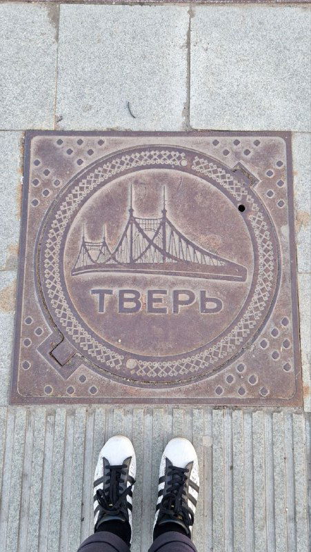 Круглый люк на тротуаре с изображением моста и надписью «Тверь», внизу кадра видны кроссовки фотографа и плитка на набережной