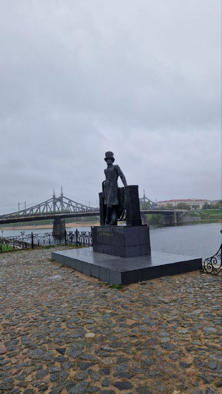 Памятник на набережной Волги в Твери, рядом виден мост через реку и булыжная набережная под серым пасмурным небом города