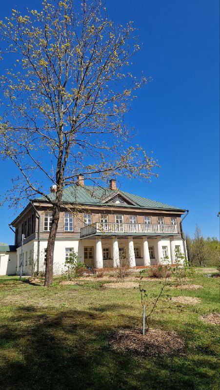 Главный двухэтажный дом усадьбы Домотканово с колоннами и зелёной крышей, окружённый газоном и молодыми деревьями в пригородном пейзаже.