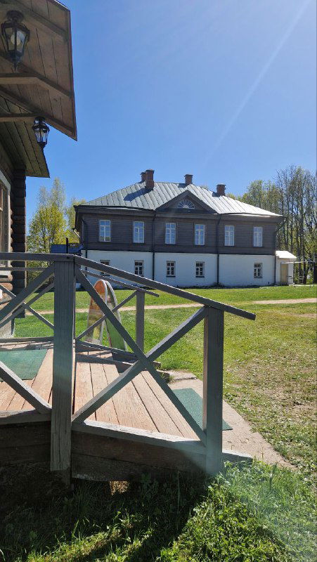 Деревянное крыльцо и перила флигеля усадьбы, вид в сторону главного дома и газона, солнечный свет и тени от деревьев весной.