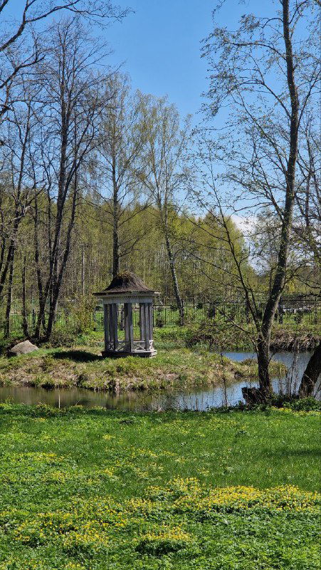 Небольшая классическая ротонда на берегу пруда в усадебном парке Домотканово, отражение в воде и весенняя зелень вокруг пруда.