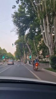 Вид из автомобиля на городскую улицу Тбилиси: машины, деревья и мопед в средней полосе, вечерняя городская сцена с движением.