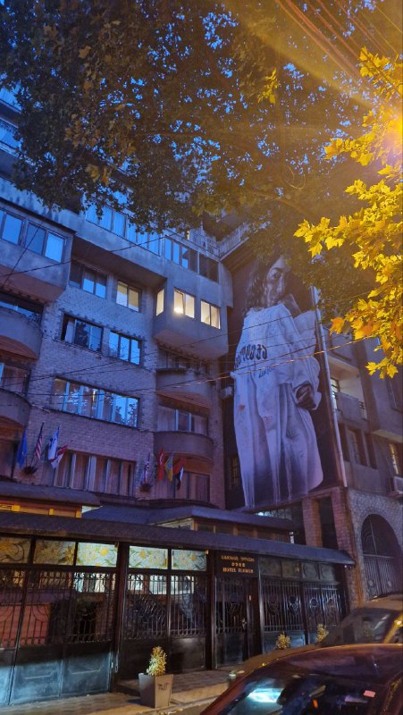 Ночная/вечерняя фотография городского пейзажа Тбилиси с домами и крупной скульптурой на фасаде — урбанистическая атмосфера центра города.