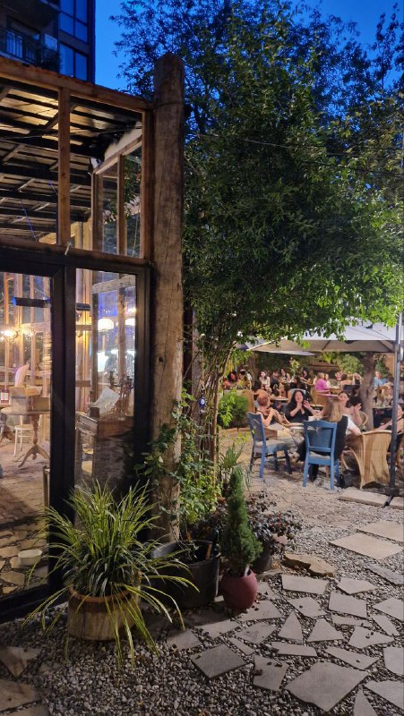 Открытая часть двора с деревянной пристройкой и столиками на каменной дорожке: много растений и люди за столами в летнем формате.