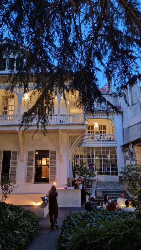 Фасад старинного дома с балконом и подсветкой, внизу видны столики на веранде — вечерний фасад заведения в тбилисском дворе.