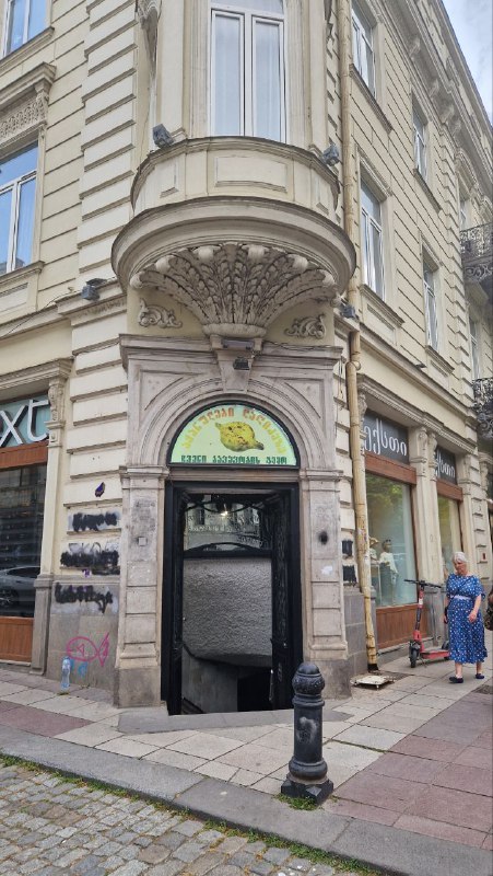 Узкий арочный вход в старинное здание с вывеской кафе и ступенями: городской фасад Тбилиси и пешеходы у входа в точку питания.