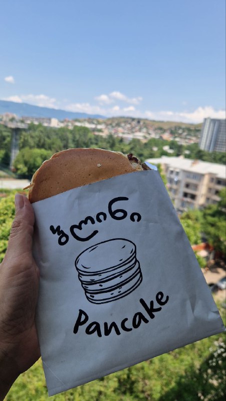 Панкейк в бумажном пакете с надписью «pancake» на фоне городской панорамы Тбилиси: уличная выпечка, купленная в гастрономе.