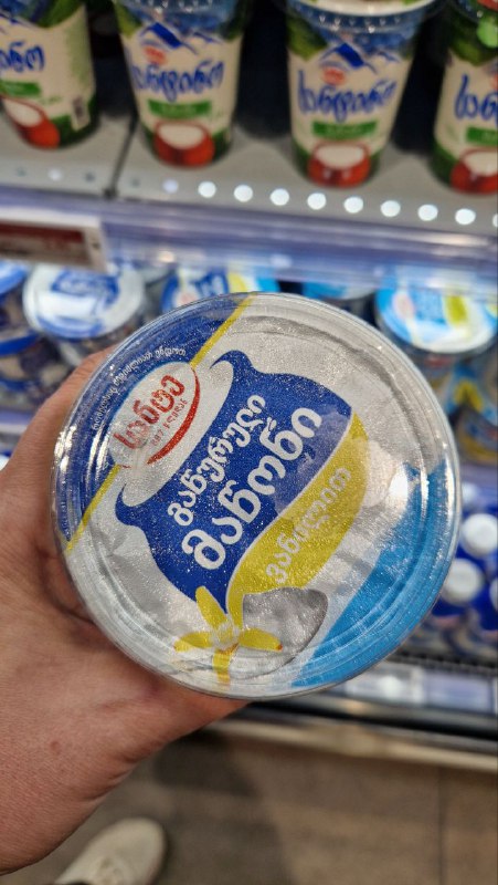 Баночка мацони с этикеткой в руке покупателя на полке магазина — демонстрация ванильного мацони и молочного ассортимента в супермаркете.