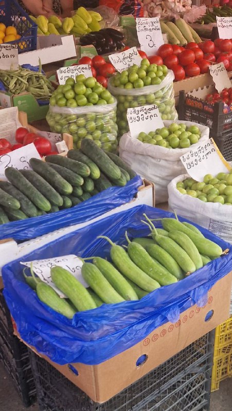 Прилавок продуктового рынка с огурцами и другими овощами в ящиках, ценниками и привычной уличной расстановкой товаров в Тбилиси.