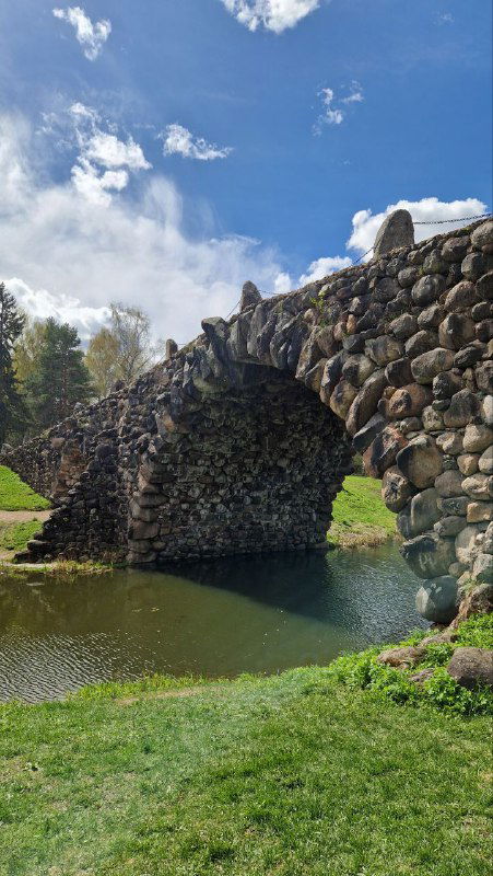 Каменный арочный мост из валунов через пруд в парковом каскаде усадьбы, отражение в воде и зелёный берег под ясным небом.