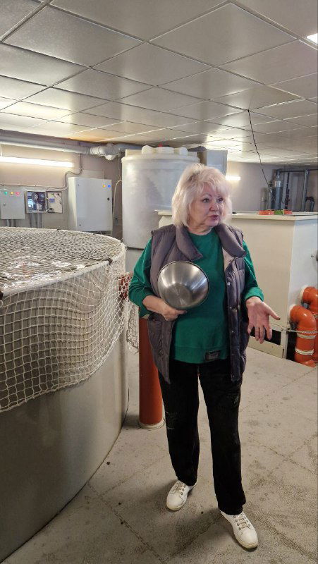 Татьяна стоит в цехе рядом с резервуарами форелевой фермы в Василёво, держит миску корма — хозяйка рассказывает о саде и местной жизни в Торжке.