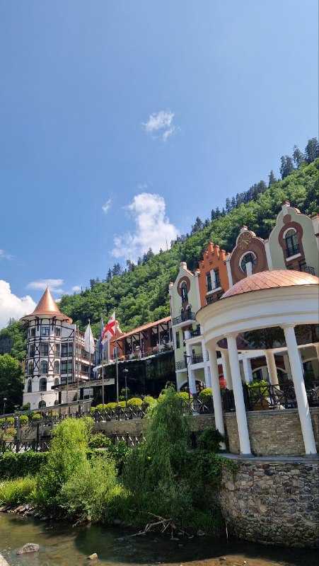 Реконструированные фасады и набережная рядом с рекой в Боржоми: цветные здания на фоне зелёного холма и ясного неба.