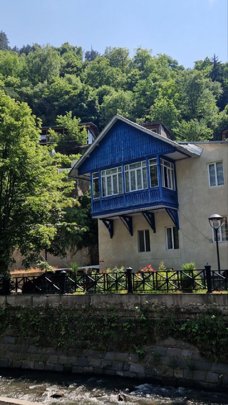Дом на склоне холма над рекой в Боржоми: голубая веранда, древесная растительность и гористый пейзаж вокруг.