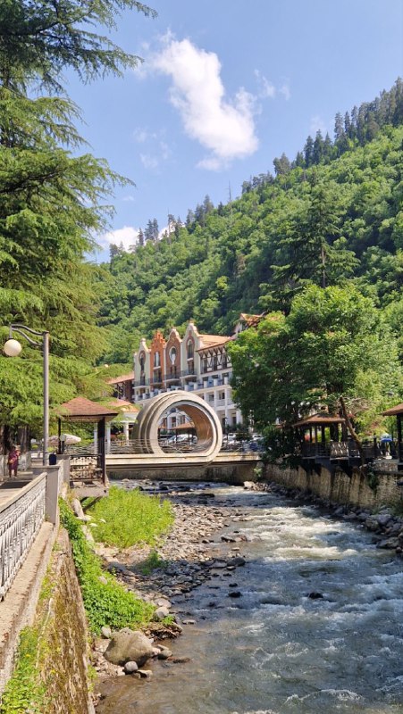 Река и мост в Боржоми: вид на водный поток, берега с зеленью и архитектурные элементы на фоне холмов и неба.