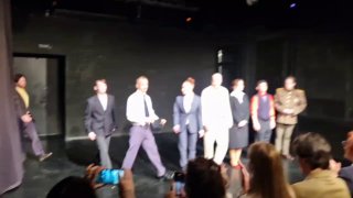 Нечёткое фото со сцены спектакля в тёмном зале: актёры на затемнённой площадке, видны зрители в первом ряду и телефон внизу кадра.