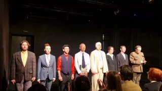 Чёткое фото актёрского состава во время поклона: ряд исполнителей в разных костюмах выстроен на тёмной сцене, зал на переднем плане.