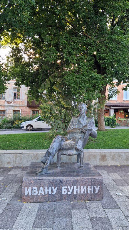 Памятник Ивану Бунину на лавочке в зелёном сквере, статуя писателя рядом с деревьями и городской застройкой на заднем плане.