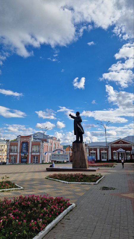 Памятник на открытой площади на фоне цветочных клумб и облачного неба, торжественный силуэт в городском ансамбле Ельца.