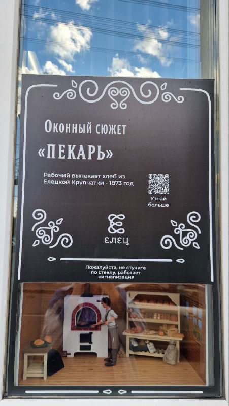 Витрина с плакатом экспозиции «Оконный сюжет «Пекарь»» и миниатюрной сценой внутри; музейная или экспозиционная витрина в городской точке.