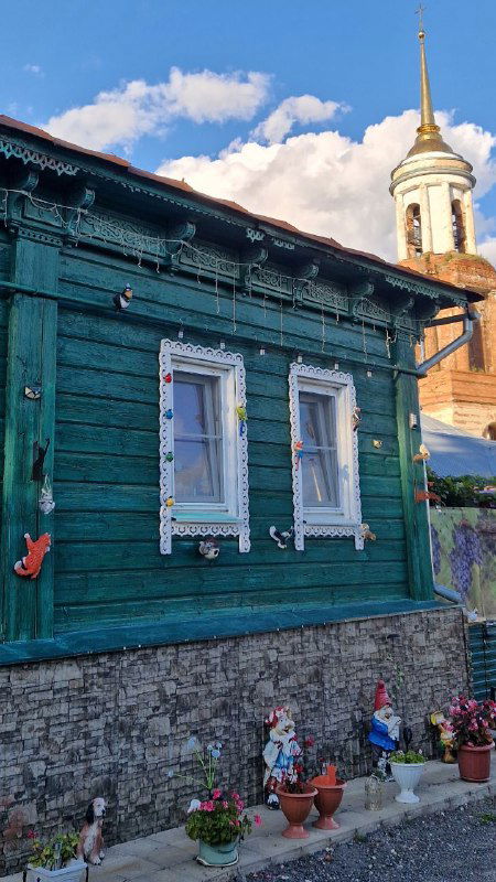 Зелёный деревянный дом с белыми наличниками на фоне церковной башни; композиция старой застройки и религиозного силуэта в Ельце.