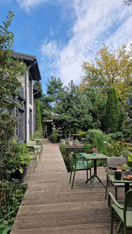 Деревянная дорожка, ведущая вдоль сада RastuS, открытая терраса с зелёными столиками и стульями и аккуратные цветочные композиции по периметру.