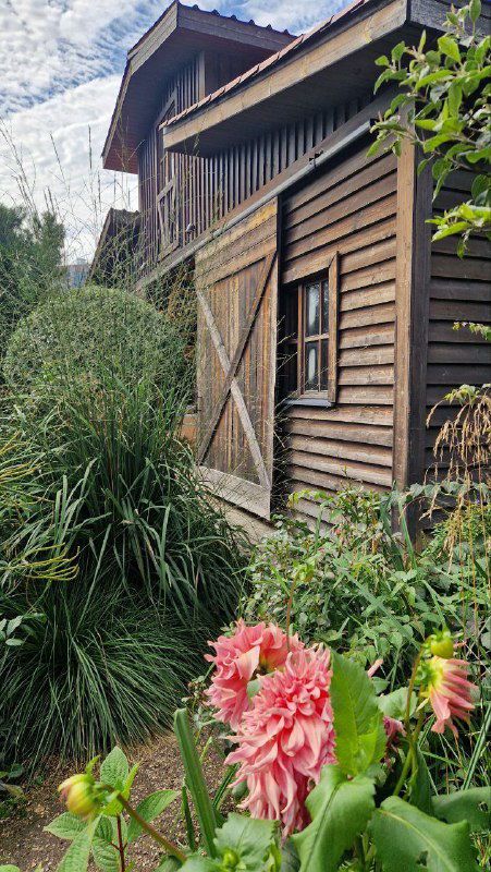 Деревянный фасад и амбарное строение в садовой части RastuS: клумбы с цветами на переднем плане и ухоженные растения вокруг входа в здание.