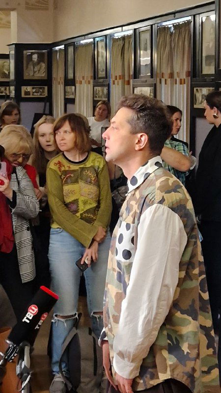 Пресс-подход: мужчина в маскировочном жилете отвечает на вопросы журналистов и публики в фойе театра у стенда спектакля.