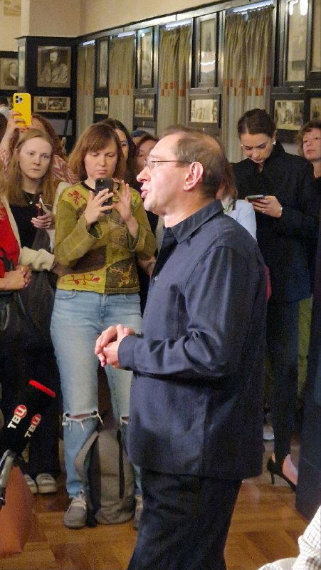 На пресс-подходе мужчина в тёмной рубашке говорит перед собравшимися журналистами и зрителями в коридоре театра.