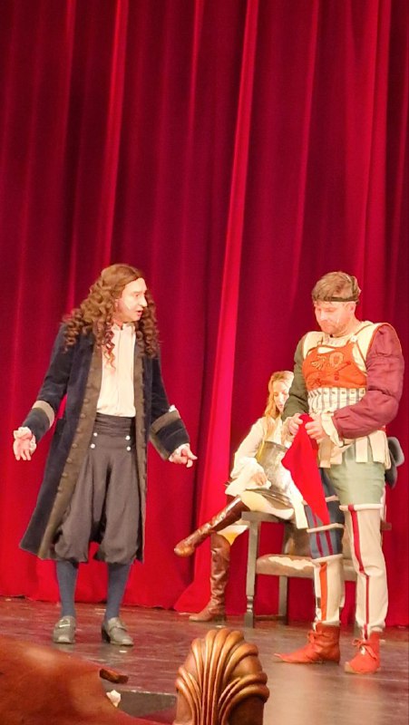 Сцена: актёры в костюмах эпохи на красном фоне занавеса, один из персонажей в длинном парике, эпизод спектакля.