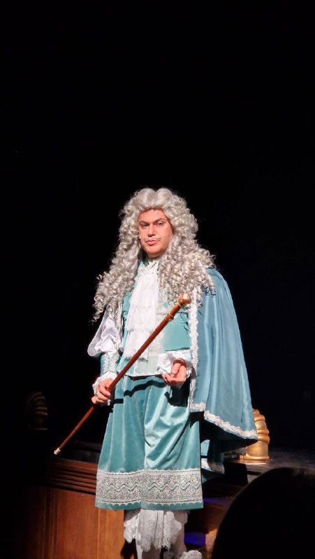 Актёр в голубом барочном костюме и парике стоит на сцене с тростью — выраженная сценическая роль и детализированный костюм.