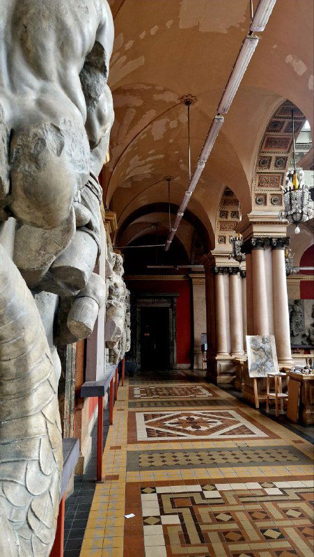 Галерея с рядами скульптур и колоннами в здании академии, видна перспектива коридора, мраморные фигуры и пол с геометрическим рисунком.