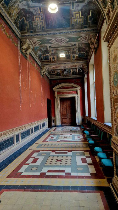 Длинный коридор академии с расписным потолком и мозаичным полом, по стенам стулья — интерьерное пространство учебных и выставочных залов.