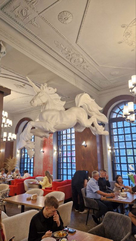 Зал с парящими скульптурами единорогов над столиками: просторное кафе с высокими окнами, посетителями и светлой атмосферой