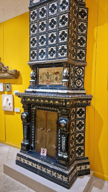 Высокая декоративная изразцовая печь в интерьере экспозиции — чёрно-белые вставки и богатый орнамент на фоне жёлтой стены музея.