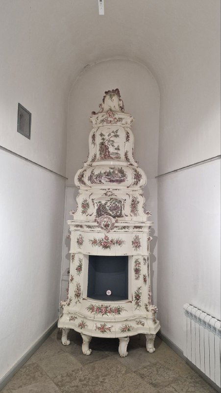 Белая старинная печь-изразцовая высокая форма в витрине — образец бытового декора прошлых столетий из собрания музея.