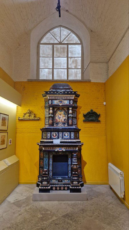 Тёмная изразцовая печь в отдельной витрине на жёлтом фоне — ещё один образец архитектурной керамики в экспозиции «Керамарх».