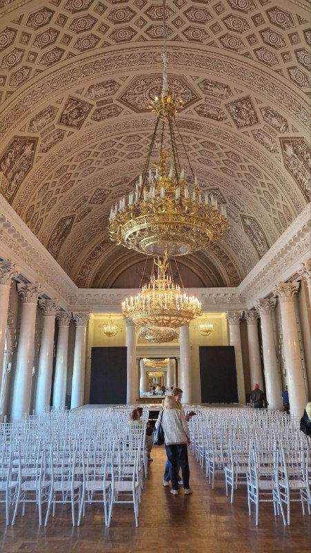 Широкий зал дворца с рядом декоративных колонн, хрустальными люстрами и белыми стульями — пространство для концертов и представлений в историческом интерьере.