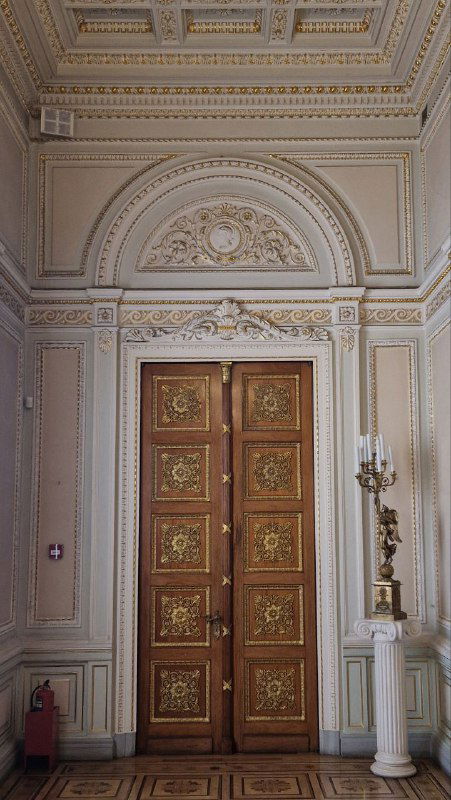 Высокие резные деревянные двери с позолоченными элементами и классическое оформление стен вокруг — парадный дверной проём во дворце.